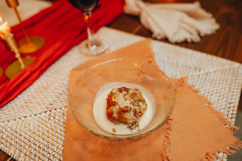 A glass bowl of dessert with a cake-like piece topped with caramel sauce, served on a rustic table setting.