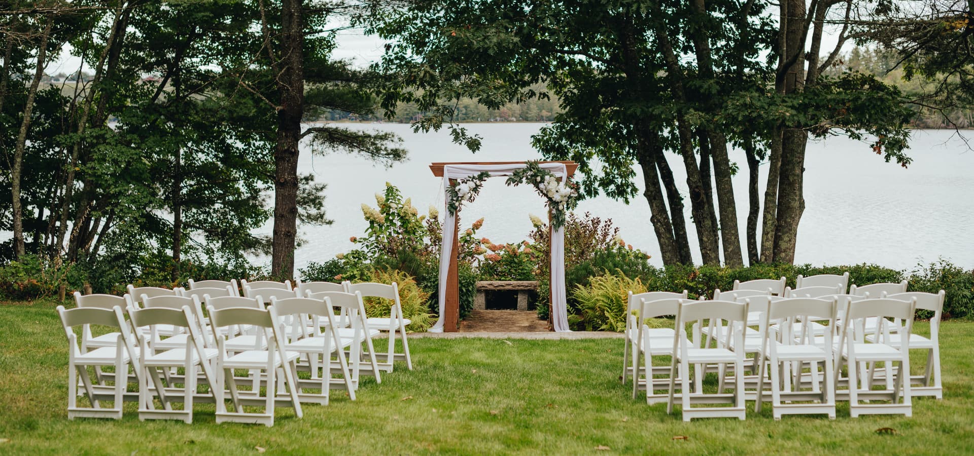 An outdoor wedding setup featuring white chairs facing a decorated arch by a serene lake.