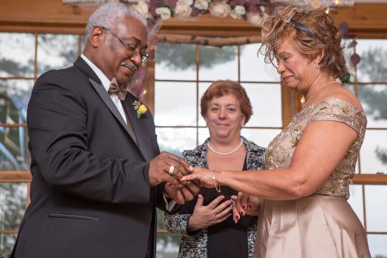 A man places a wedding ring on a woman's finger during a ceremony, while a witness stands behind them.