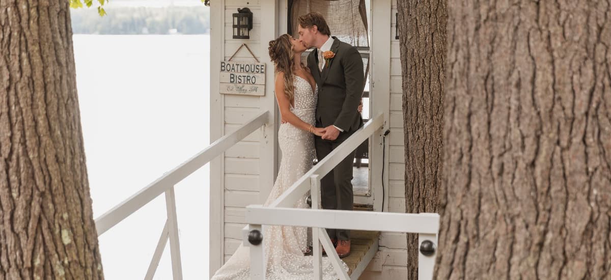 A couple kisses on a dock in front of a charming boathouse.