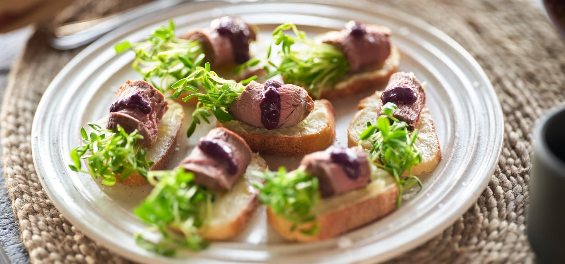 A plate of bread slices topped with beef, greens, and a berry sauce.