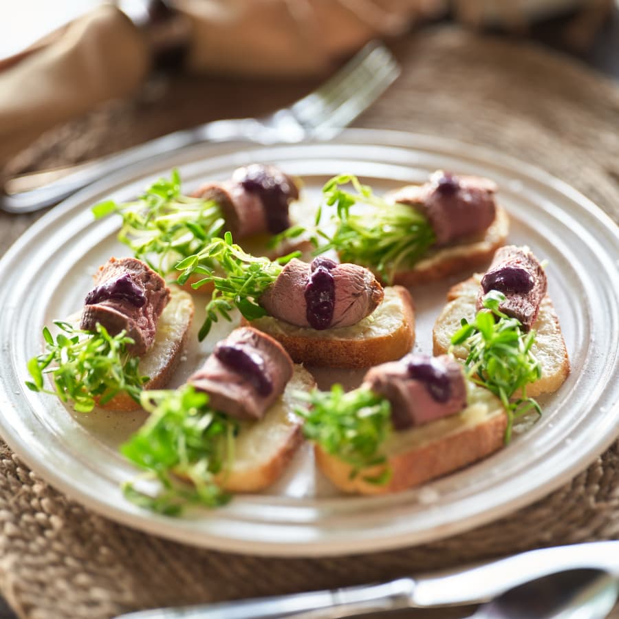 A plate of crostini topped with sliced meat, greens, and a drizzle of sauce.