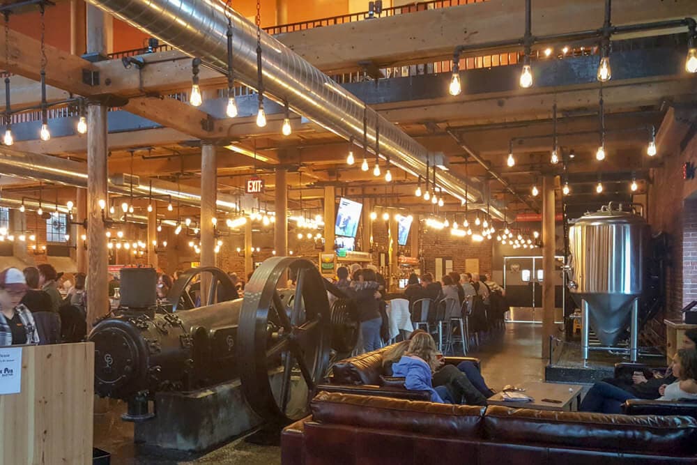 A spacious industrial-style bar filled with patrons, featuring exposed brick, vintage machinery, and warm pendant lighting.