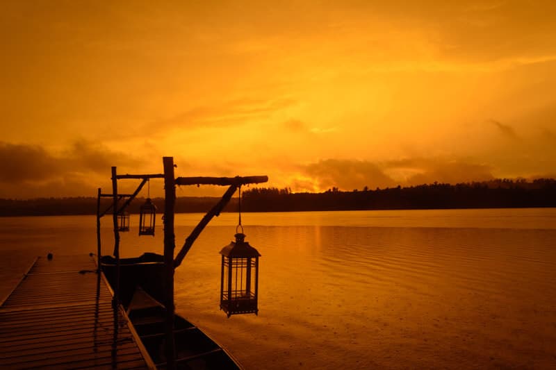 Silhouetted lanterns hang from a wooden dock against a striking orange sunset over a calm lake.