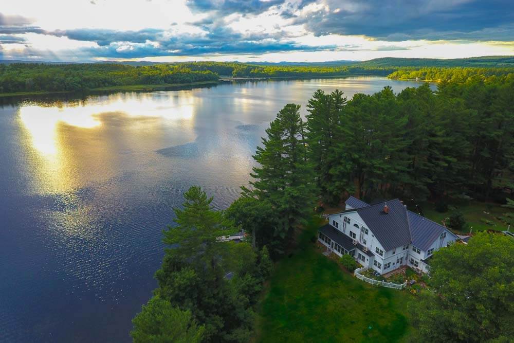 A serene lake scene with a house surrounded by trees under a cloudy sky.