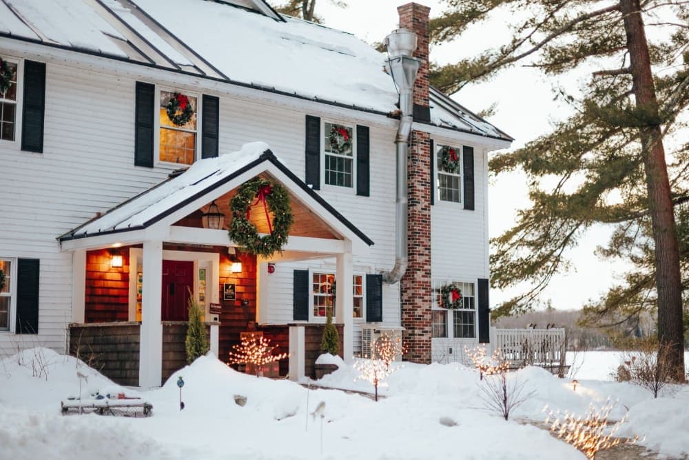 A cozy white house decorated with holiday wreaths, surrounded by snow and lit with warm outdoor lights.