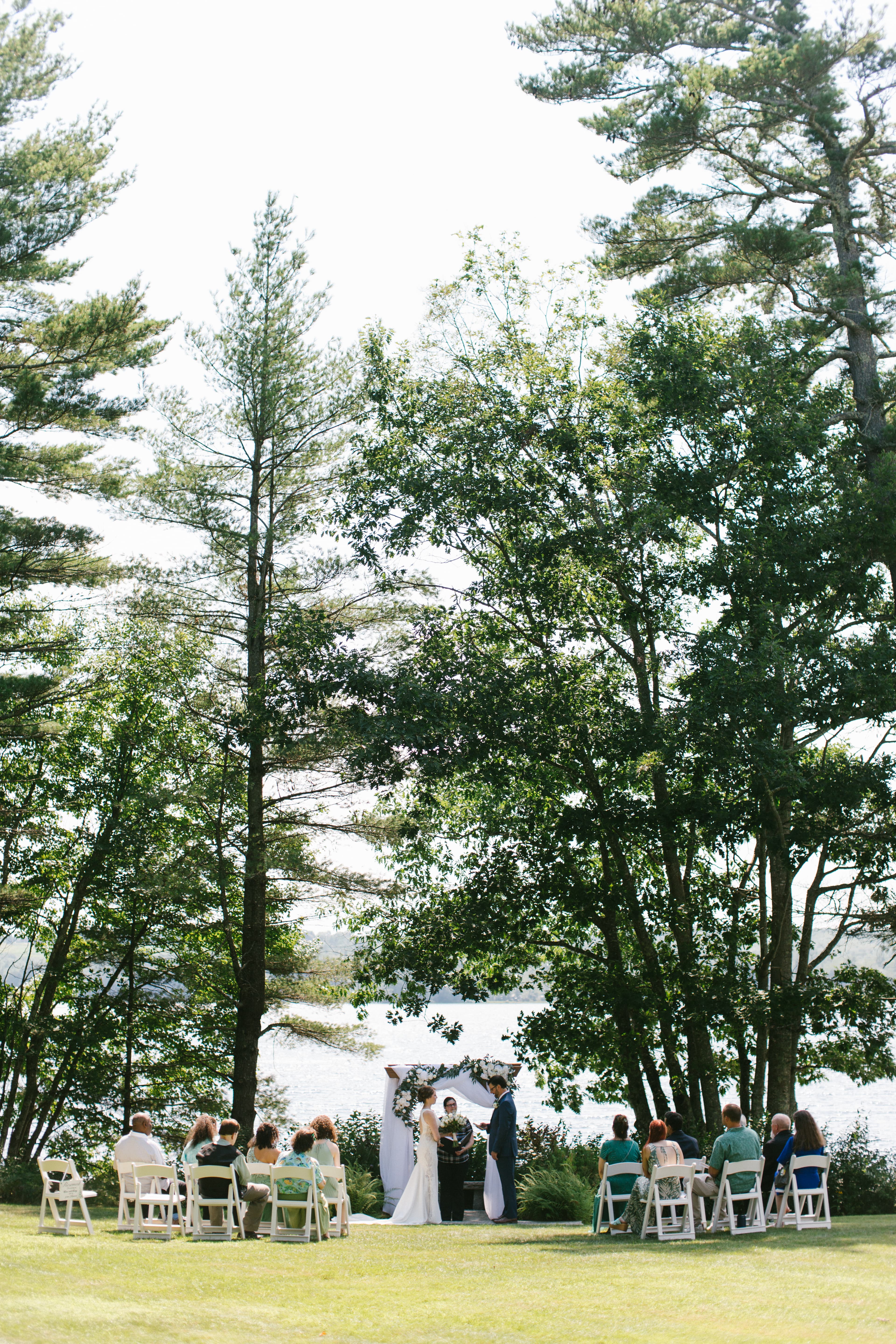 A couple exchanges vows under a floral arch by a lake, surrounded by guests seated on white chairs.