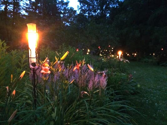 A garden pathway illuminated by glowing torches at dusk, surrounded by blooming flowers.