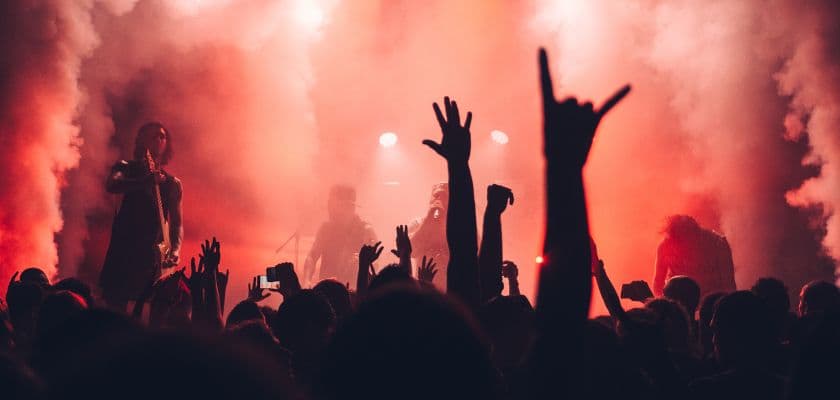 A concert scene with silhouetted fans raising their hands amidst colorful smoke and stage lights.