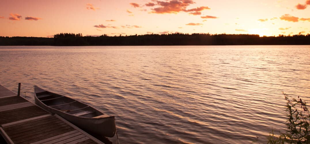 A calm lake at sunset reflects pastel colors, with a canoe resting on a dock.