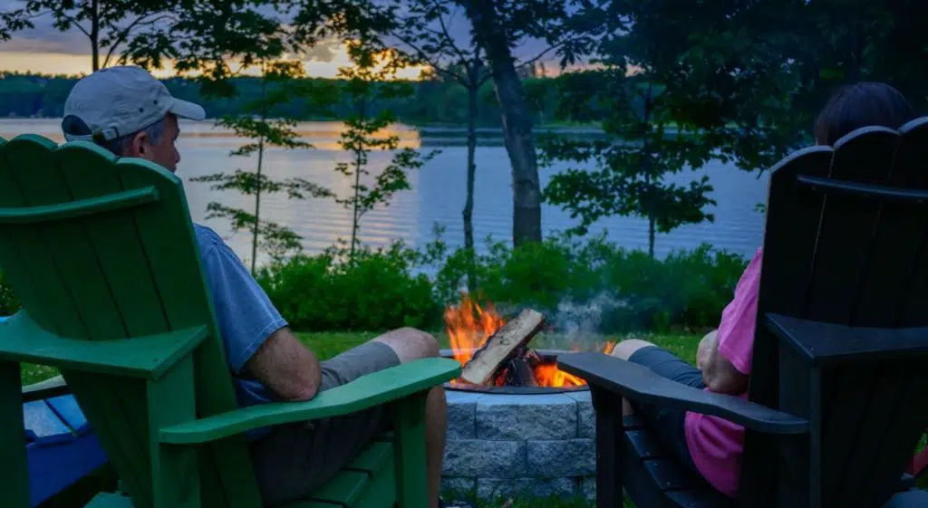 A couple sits by a campfire on a serene lakeside at sunset.