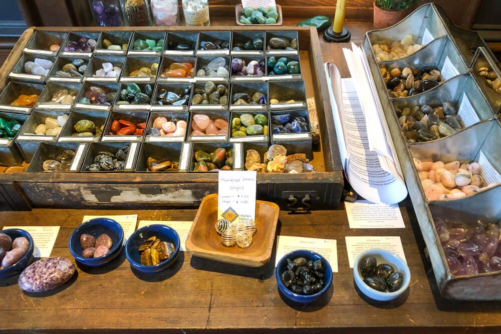 Display of various gemstones and crystals in trays and bowls on a wooden table.