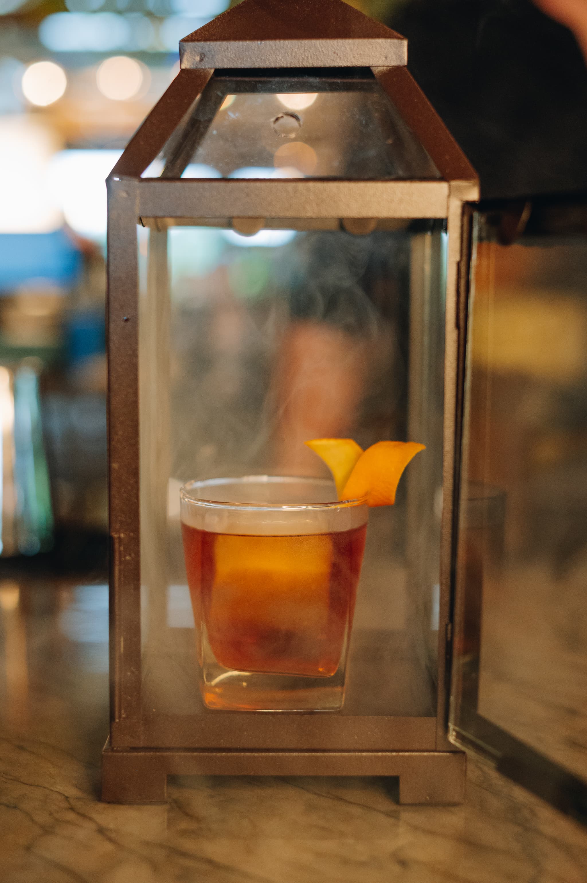 A cocktail glass with orange peel is placed inside a glass lantern, emitting a swirl of smoke.