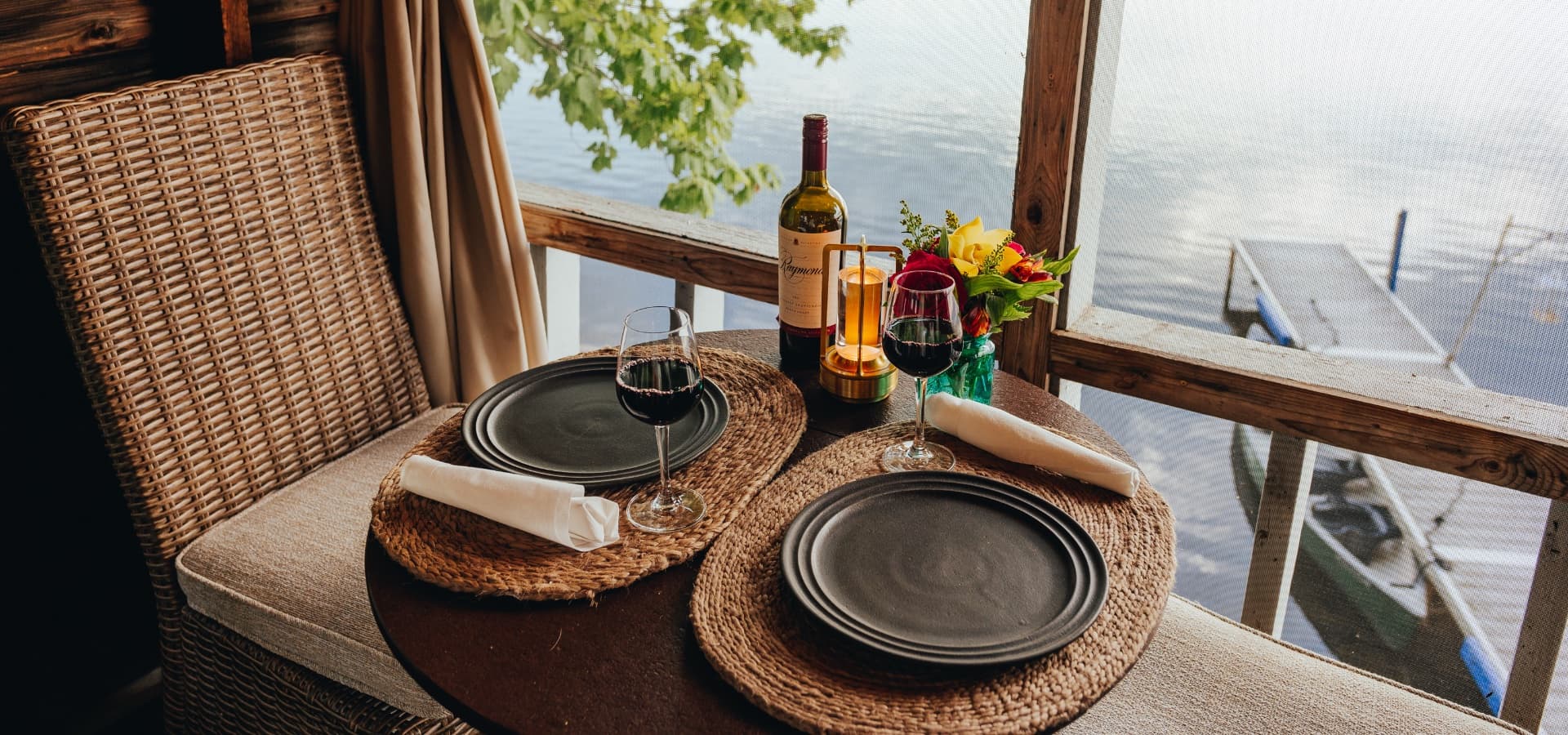 A cozy dining table by the water, set for two with wine glasses, a bottle of wine, and a colorful floral arrangement.