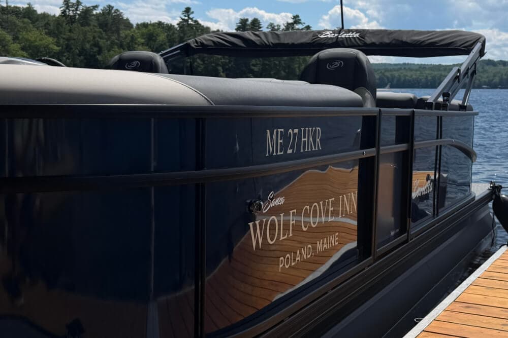 A black boat with "Wolf Cove Inn" and "Poland, Maine" printed on its side, docked by a lakeside.