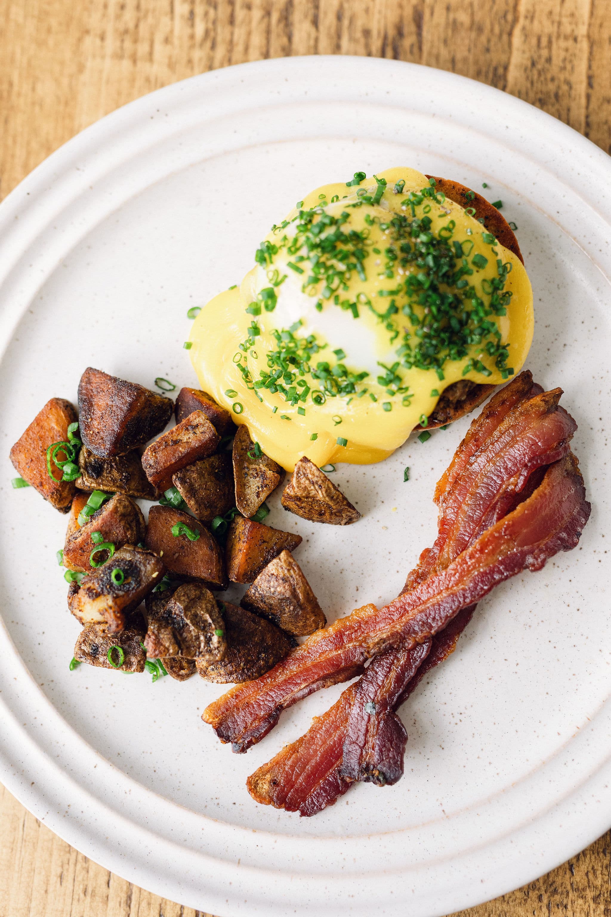 A plate featuring eggs Benedict topped with hollandaise sauce and chives, accompanied by roasted potatoes and strips of bacon.