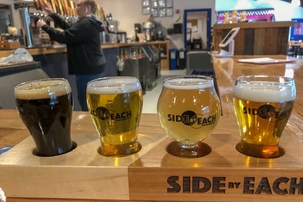 A flight of four beer samples on a wooden tray in a brewery setting.