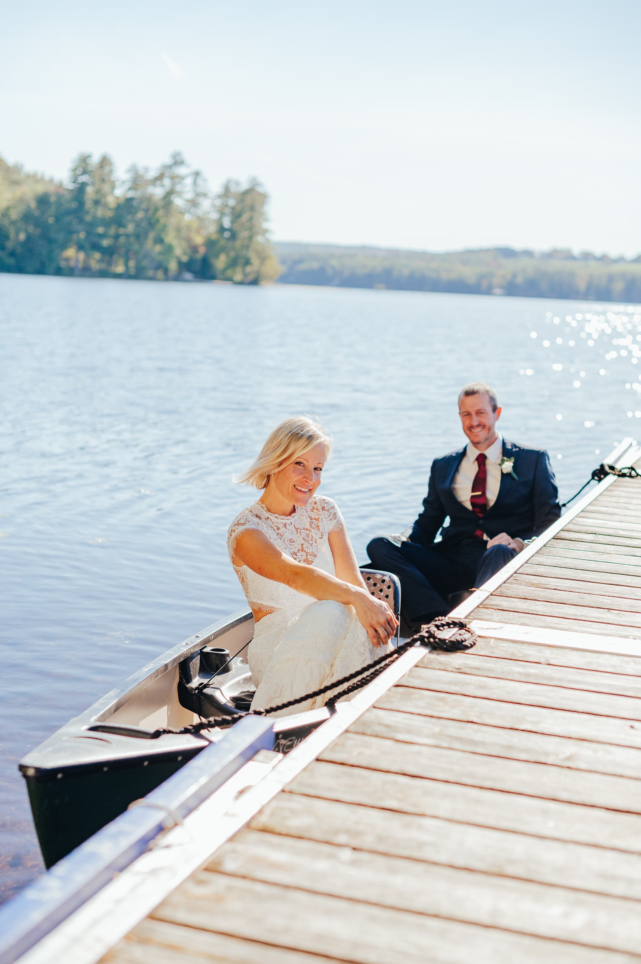 A bride and groom sit side by side in a canoe near a wooden dock, surrounded by a serene lake.
