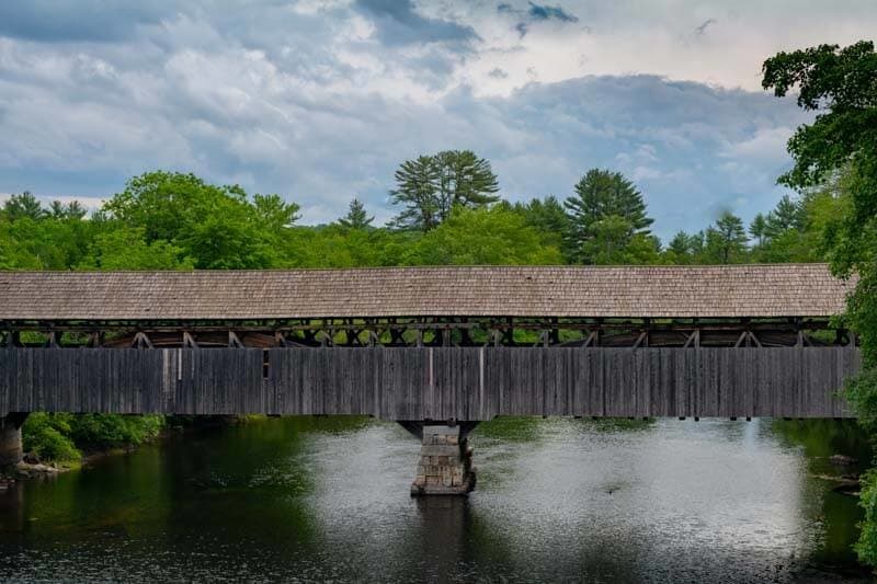 A wooden covered bridge spans a calm river surrounded by lush green trees under a cloudy sky.