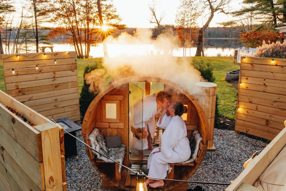 A couple in white robes kisses inside a steaming wooden sauna at sunset by a lake.