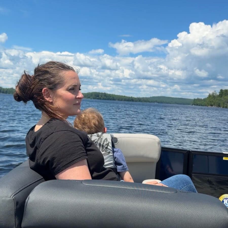 A woman sits on a boat by a lake, gazing at the water while holding a child.