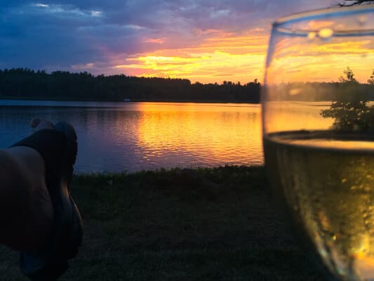 A glass of white wine in the foreground with a sunset reflecting on a lake in the background.