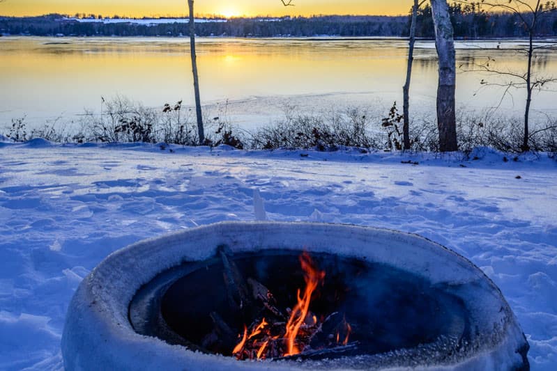 A fire burns in a circular pit with a snowy landscape and a sunset reflecting on a lake in the background.