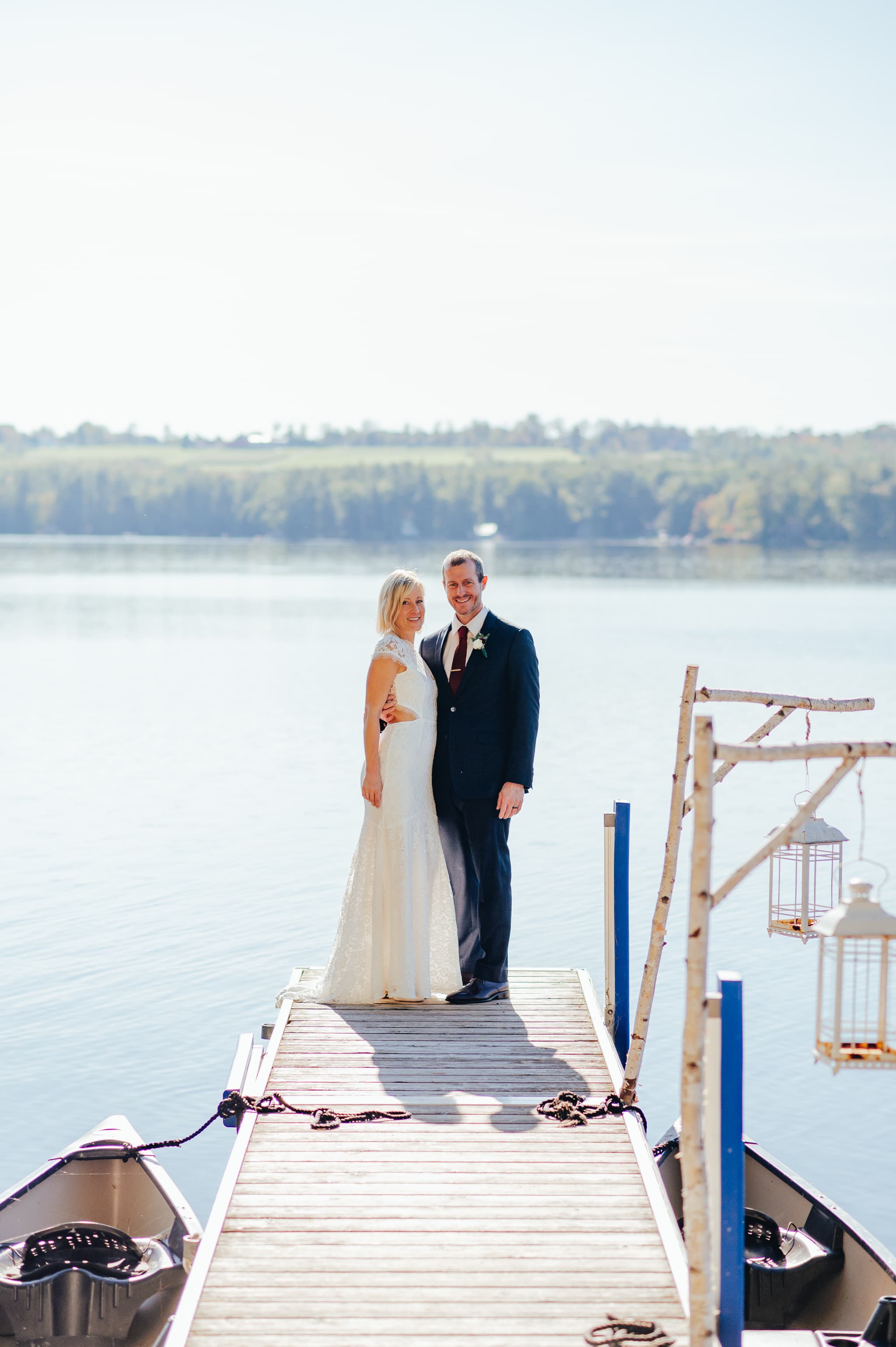 A bride and groom stand together on a dock overlooking a serene lake.