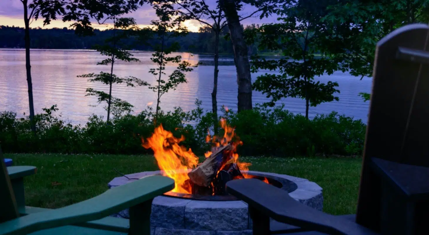 A fire burns in a stone pit overlooking a serene lake at sunset.