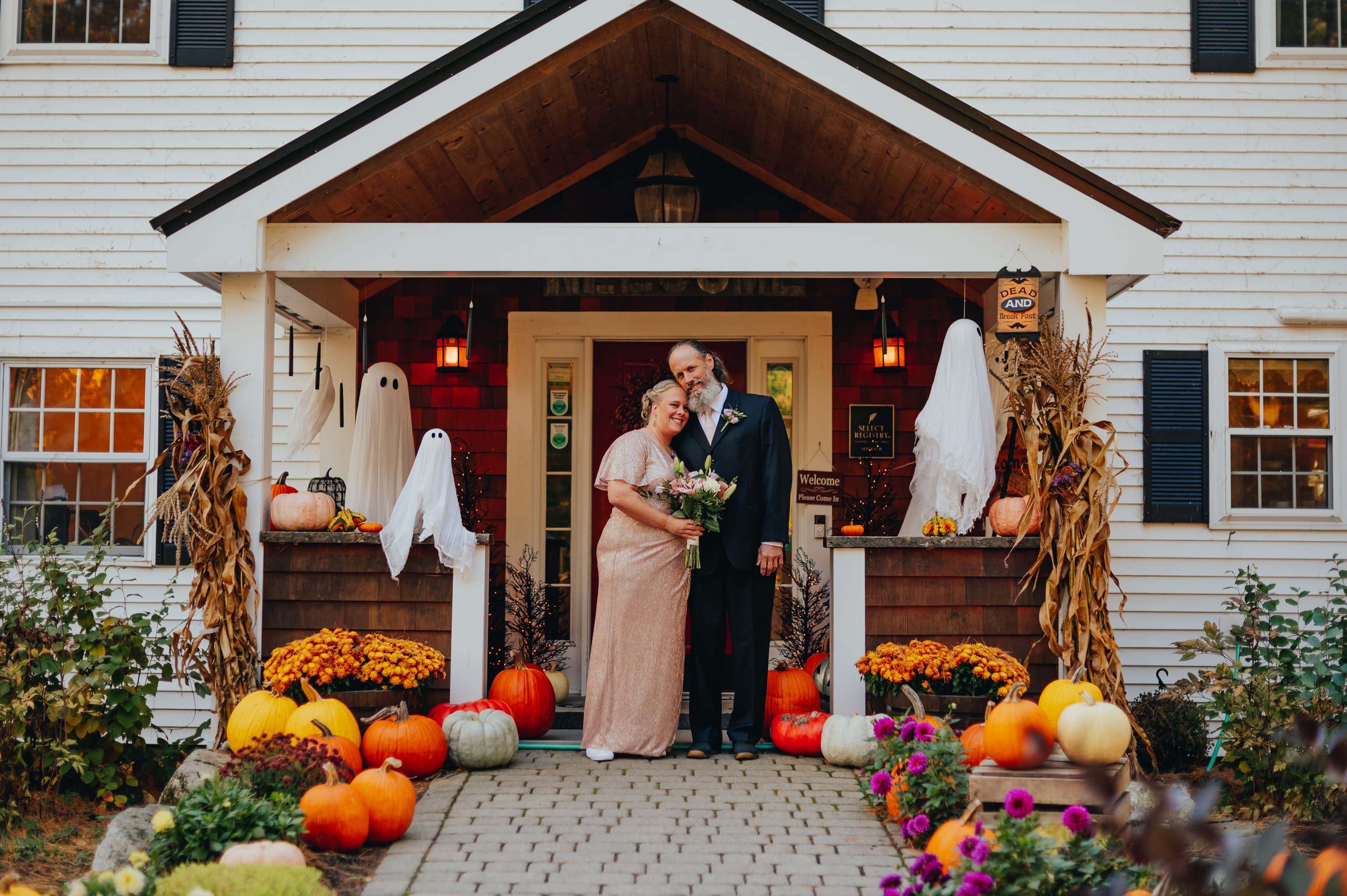 A couple smiles together on a decorated porch with pumpkins and Halloween decorations.