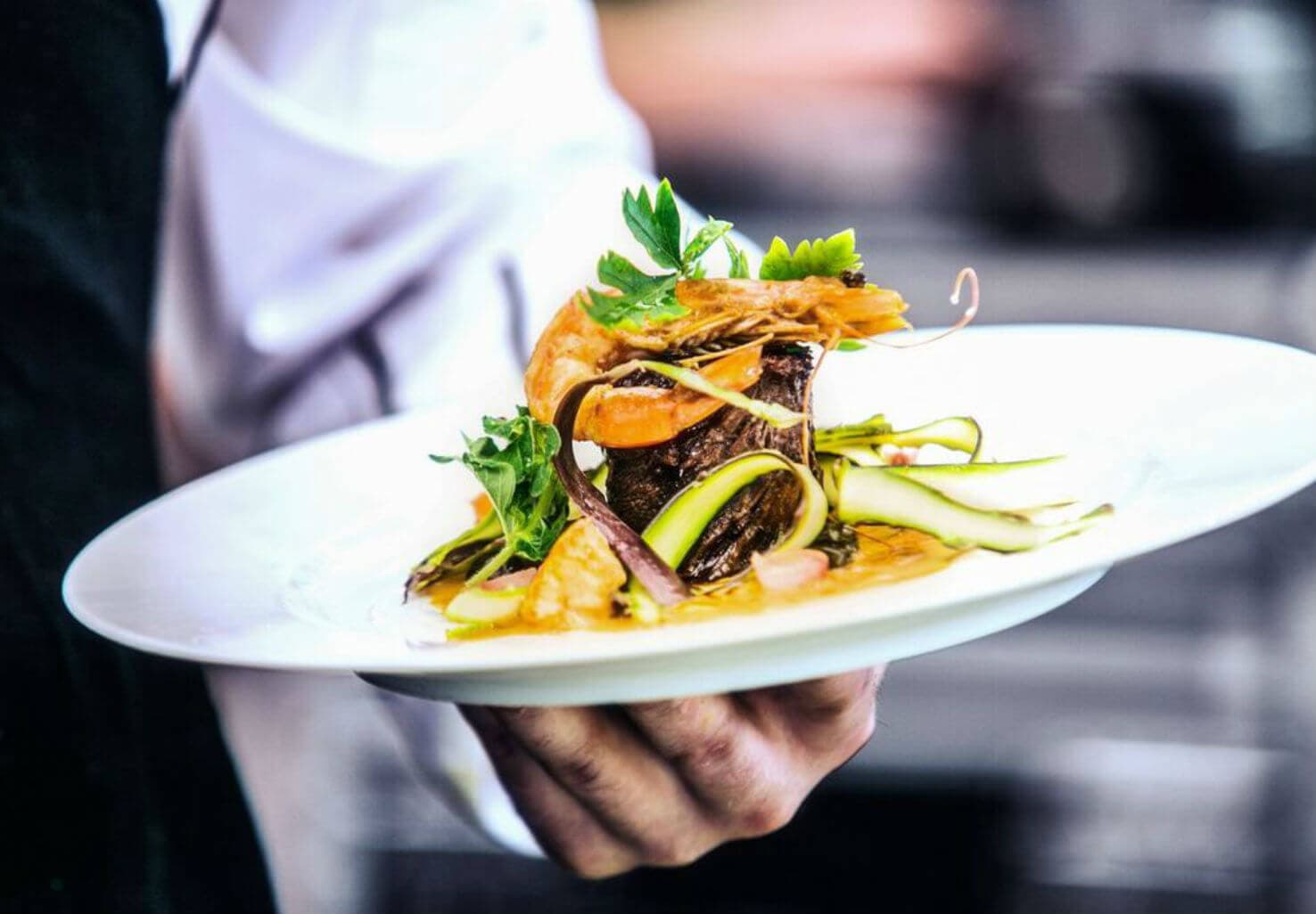 A chef presents a beautifully plated dish featuring meat, seafood, and fresh vegetables.