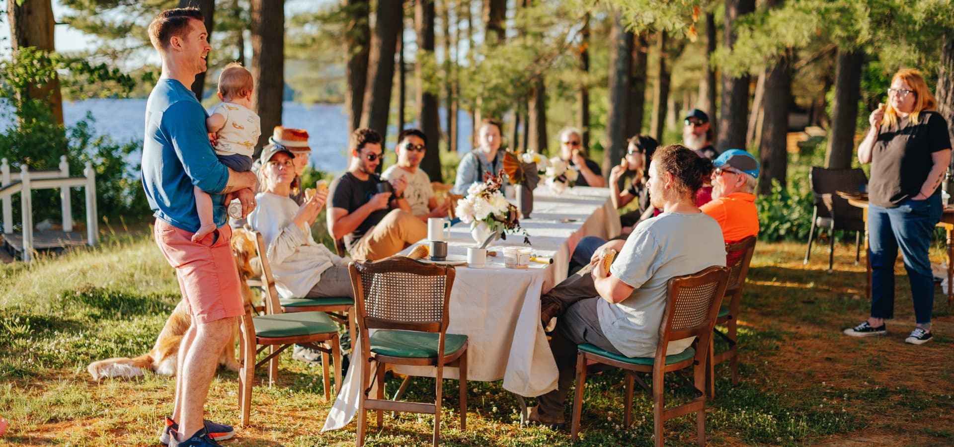 A group of people enjoys a meal together outdoors by a lake, with trees in the background.