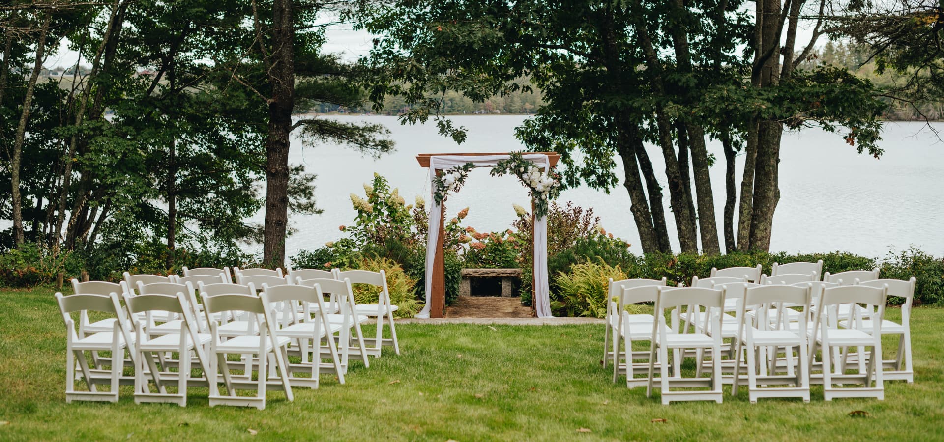 A serene outdoor wedding setup featuring white chairs facing an adorned arch by a lake, surrounded by lush greenery.