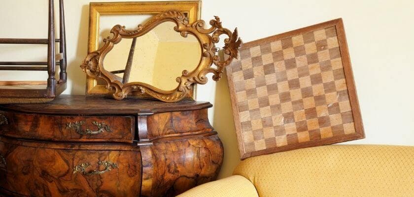 An ornate mirror and a wooden chessboard lean against a vintage dresser near a yellow sofa.