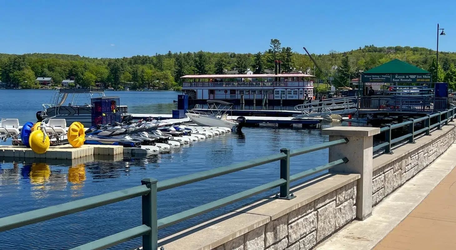 A scenic view of a marina with parked jet skis and a boat docked on a calm lake surrounded by greenery.