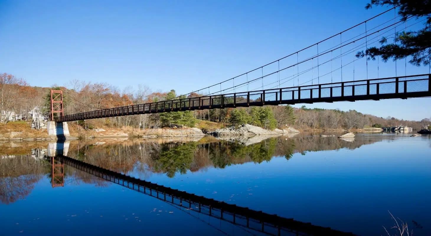 A suspension bridge spanning a calm river with reflections of trees and rocks on the water's surface.