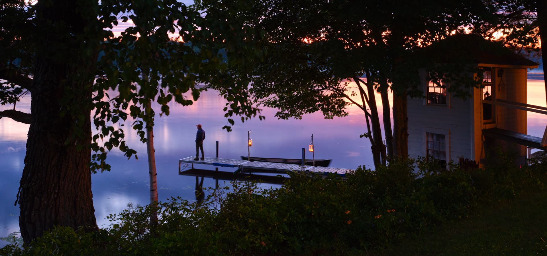 A person stands on a wooden dock by a calm lake at sunset, surrounded by trees.
