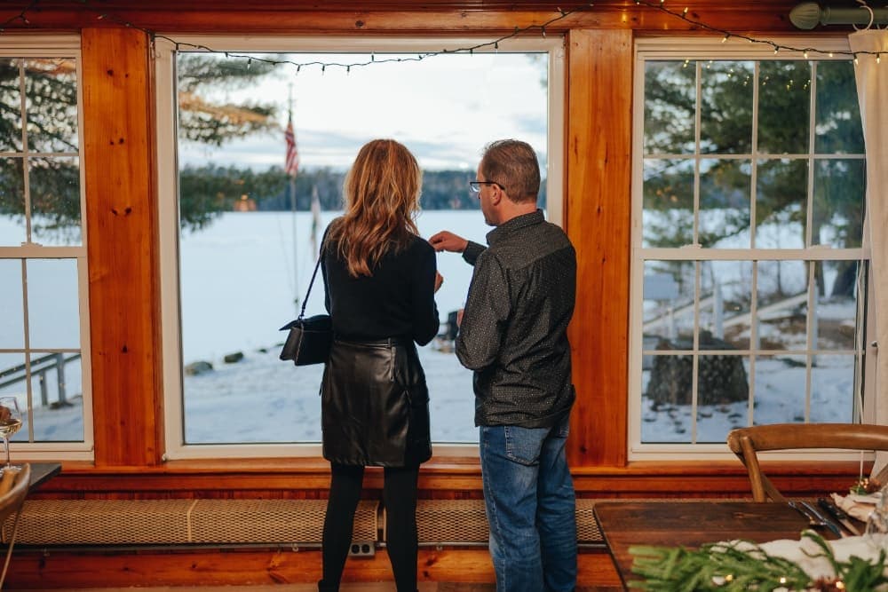 A couple stands by a window, gazing at the snowy landscape outside.