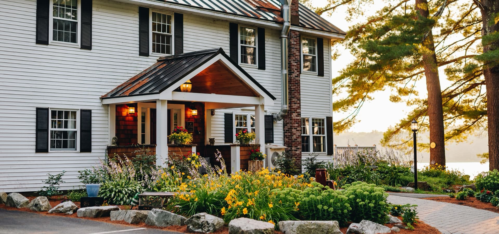 Charming white house with a porch surrounded by colorful flowers and greenery by the water.