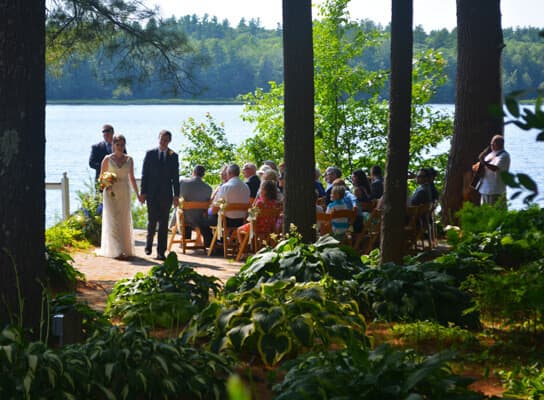 A bride and groom walk towards a seated audience by a lake, surrounded by greenery.
