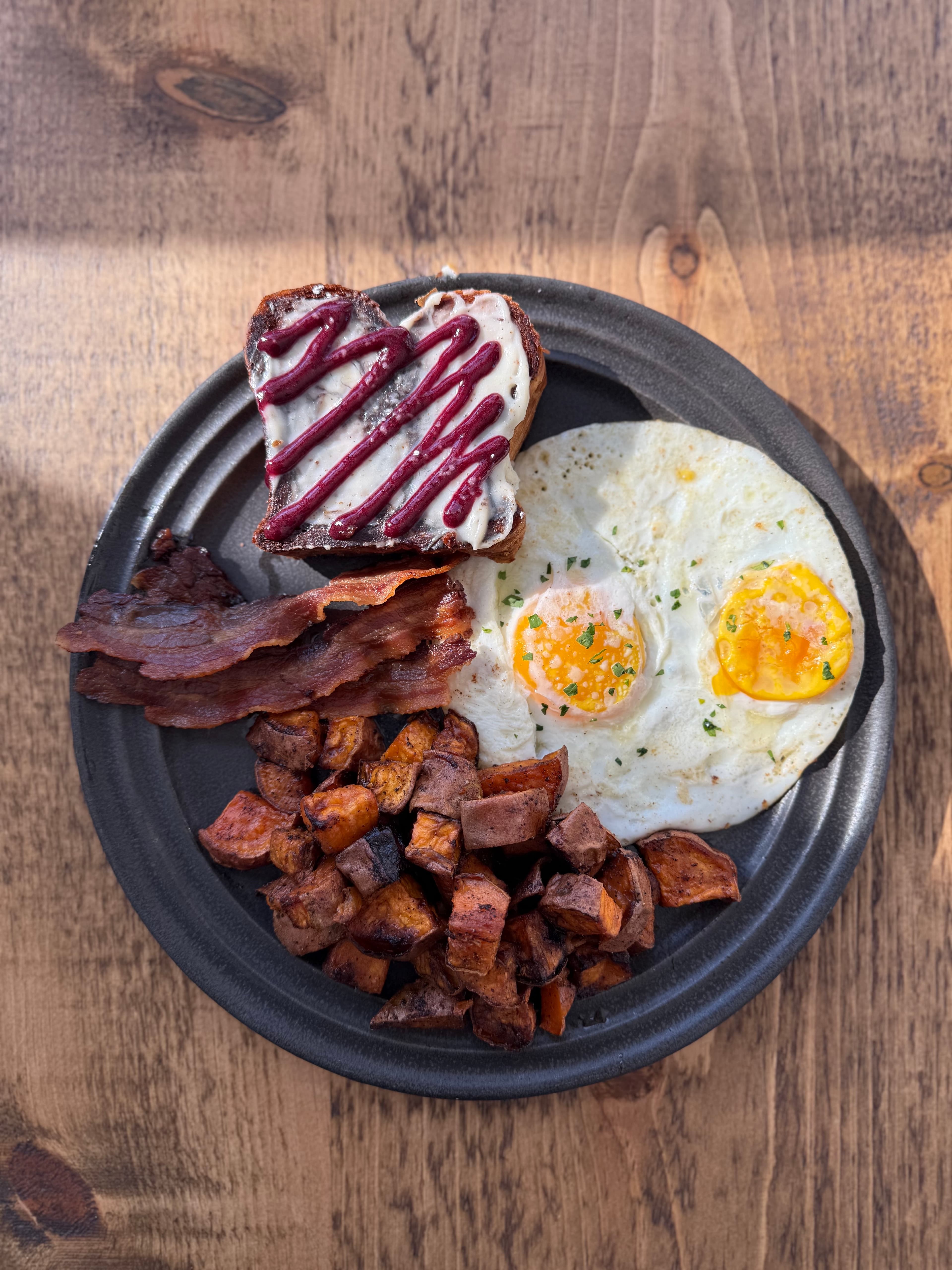 A plate of breakfast featuring two fried eggs, crispy bacon, sweet potato cubes, and a slice of toast topped with cream and berry drizzle.