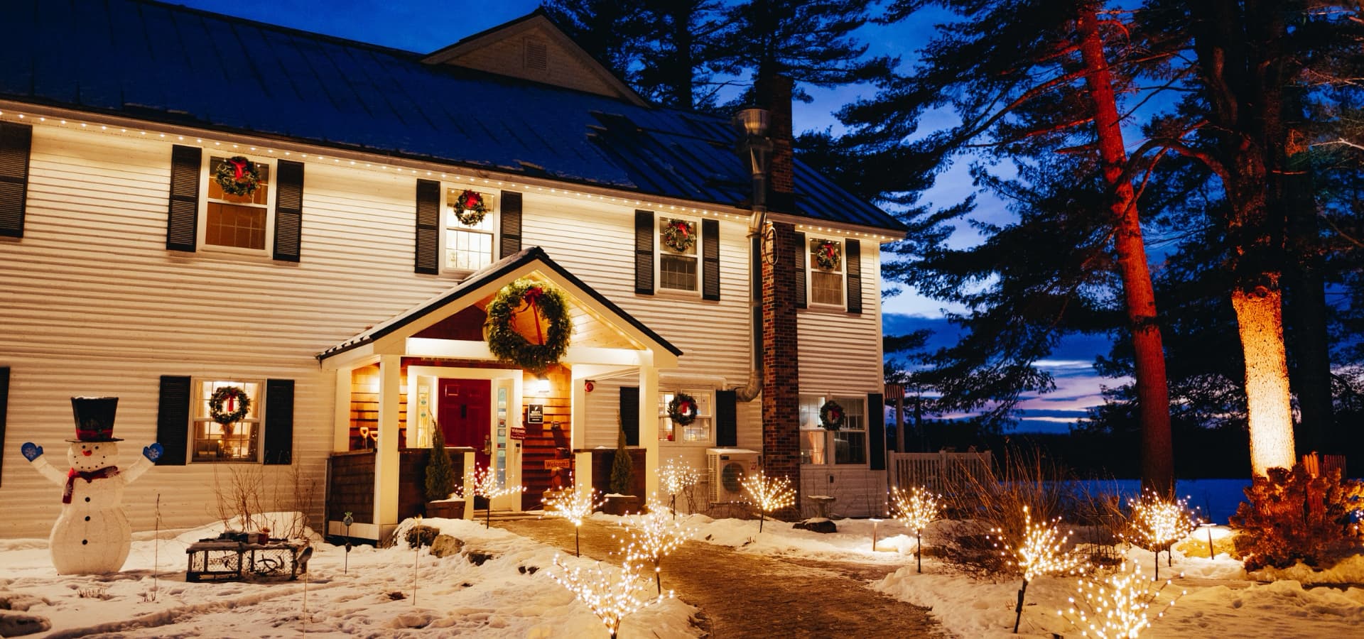 A cozy, snow-covered house adorned with Christmas decorations and illuminated by warm lights at dusk.