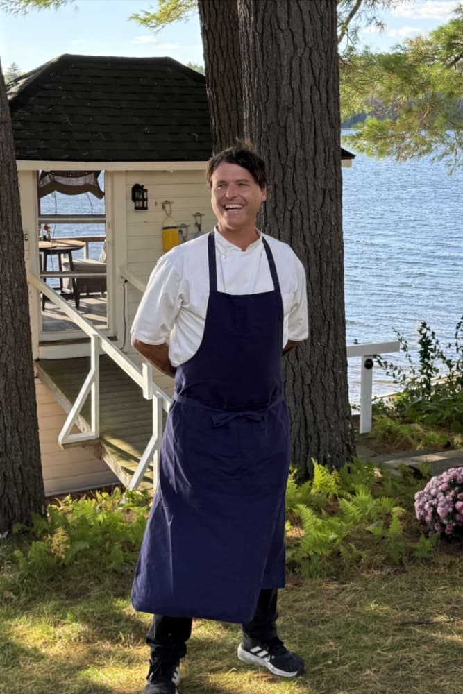 A smiling chef in a blue apron stands outdoors by a serene lake setting.