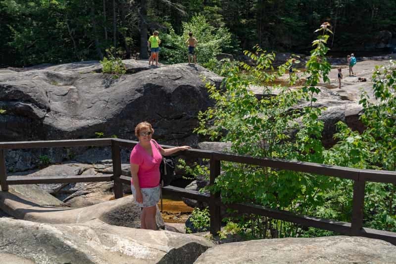 A woman in a pink shirt stands on a wooden path surrounded by greenery and large rocks, with people visible in the background.