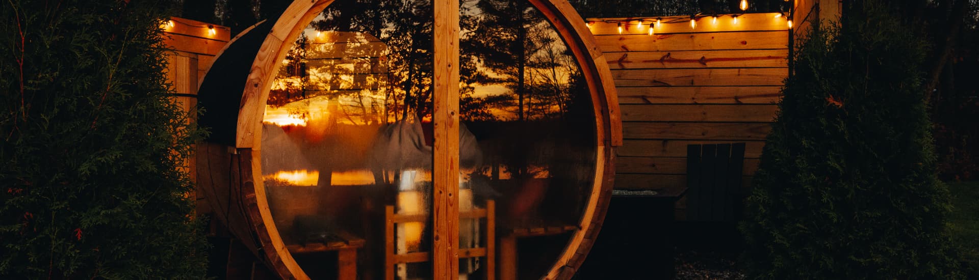 A circular wooden structure with a glass front reflects a colorful sunset, surrounded by greenery and warm lights.