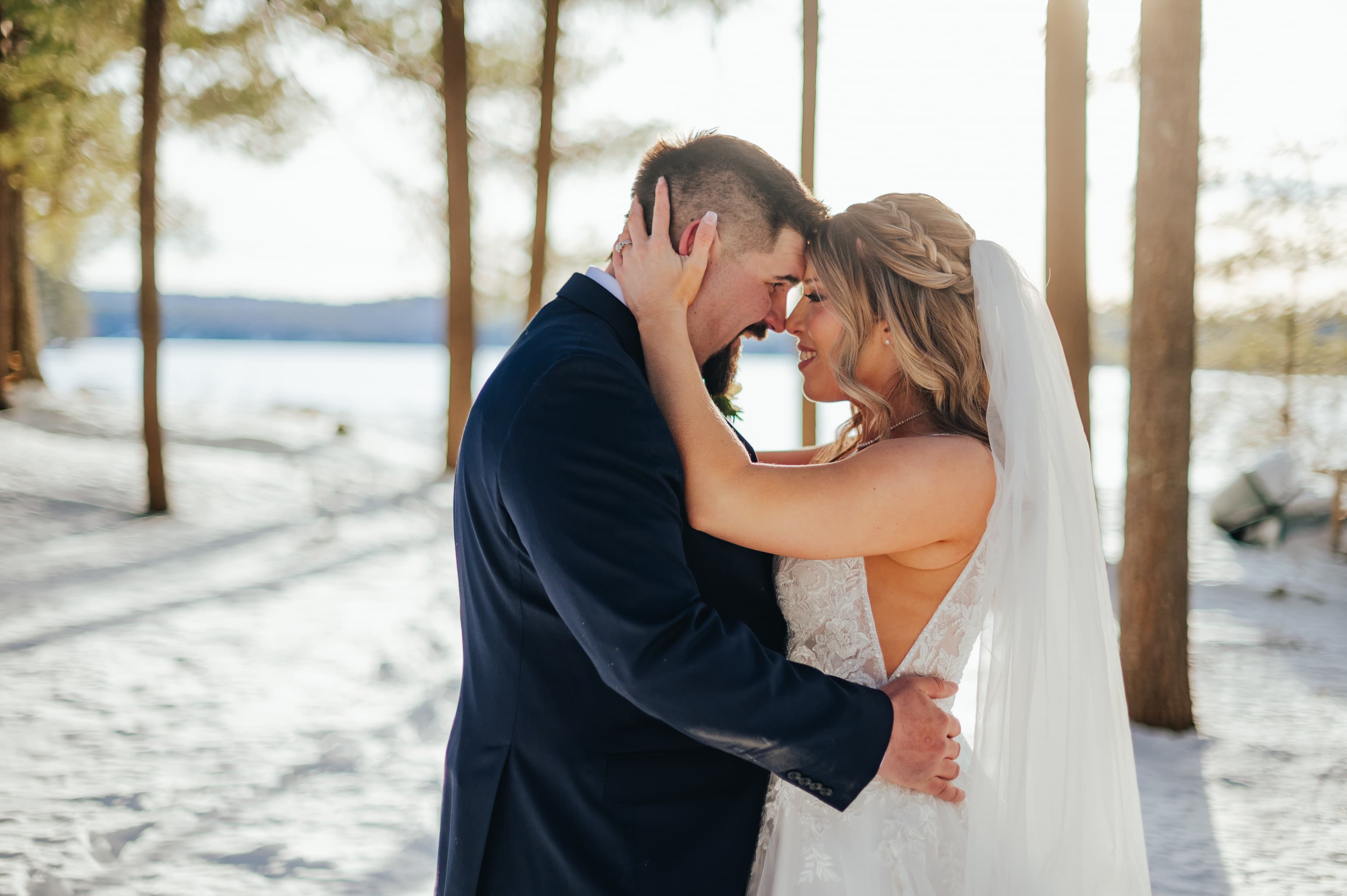 A couple embraces tenderly in a snowy landscape, framed by tall trees and a serene body of water in the background.