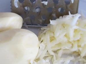 Peeled potatoes next to freshly grated potato shreds and a grater.