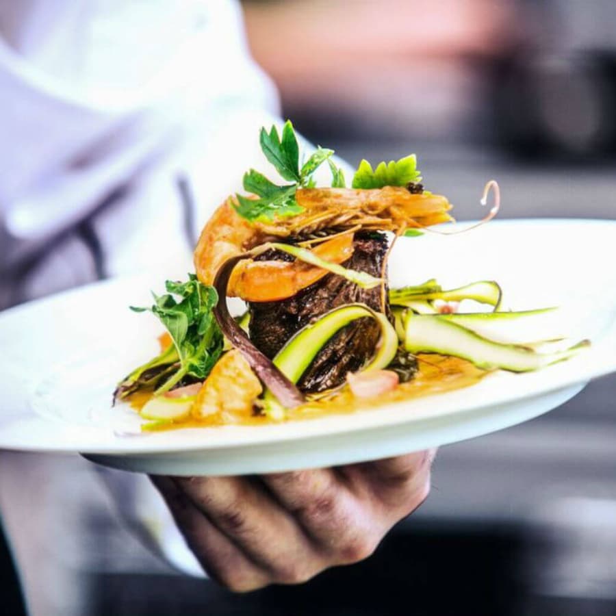 A chef presents a beautifully plated dish featuring shrimp, beef, and fresh vegetables.