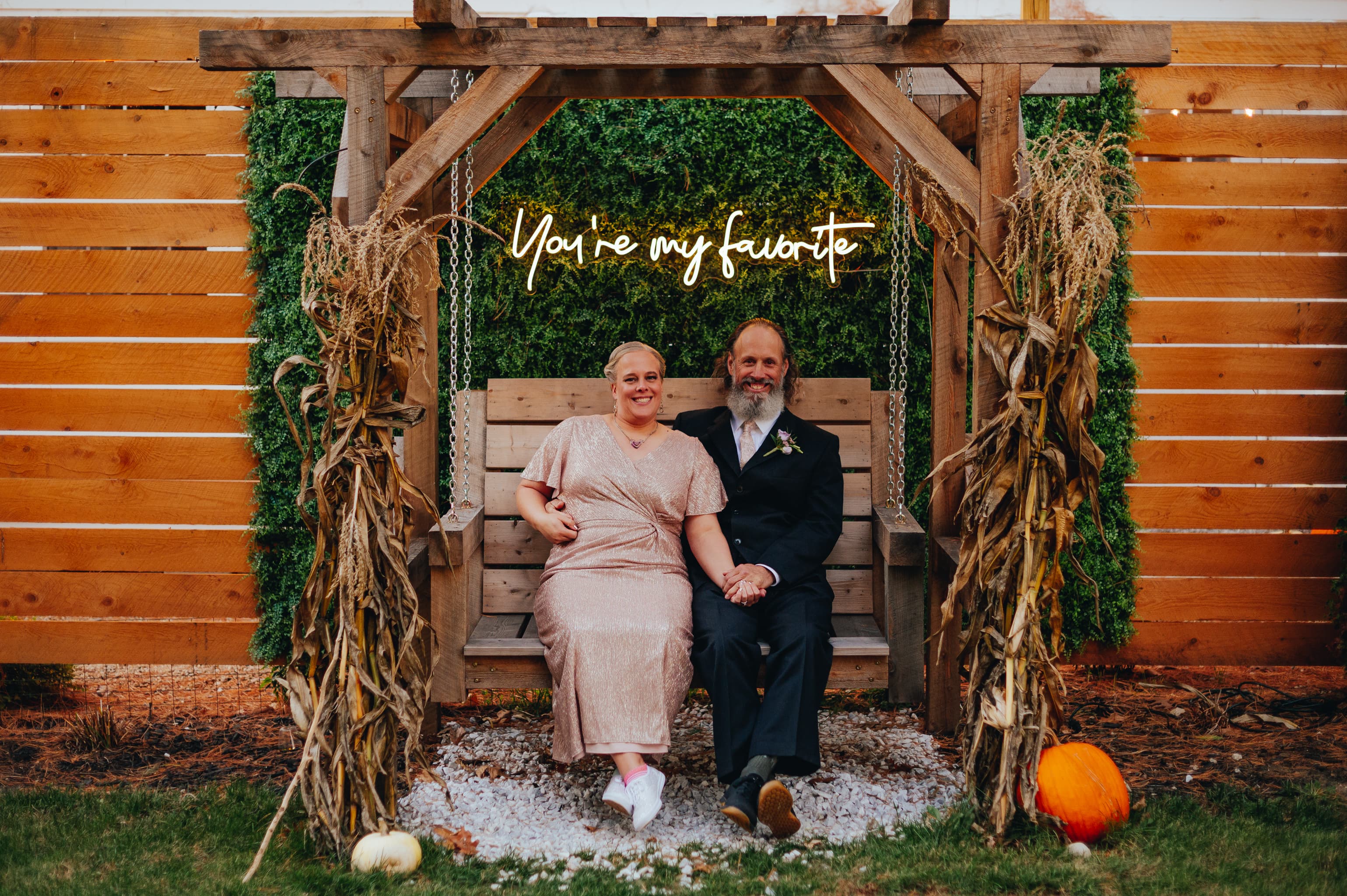 A couple sits on a wooden swing surrounded by greenery and autumn decor, with a sign reading "You're my favorite" above them.