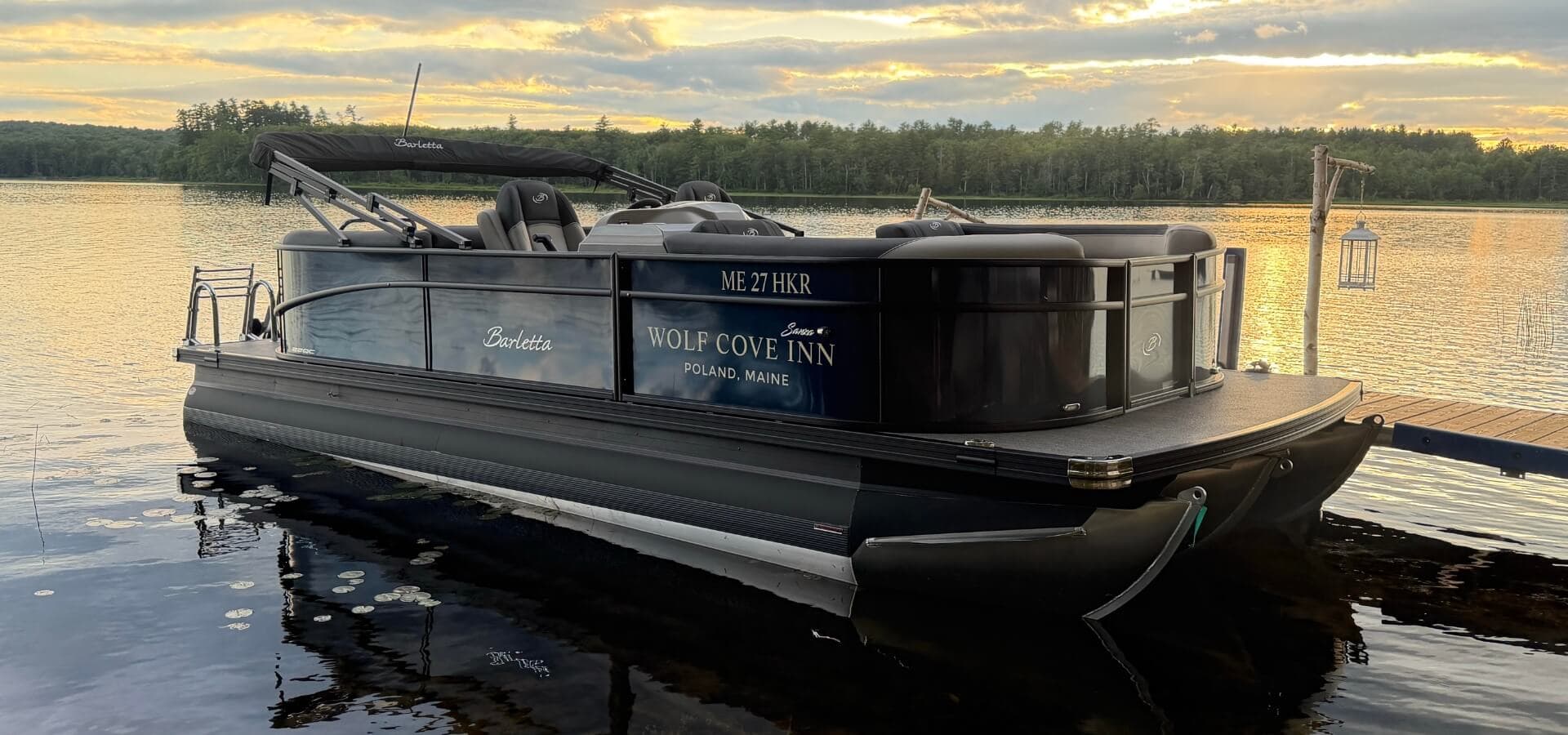 A black pontoon boat named "Scarlett" docked in a calm lake during sunset.